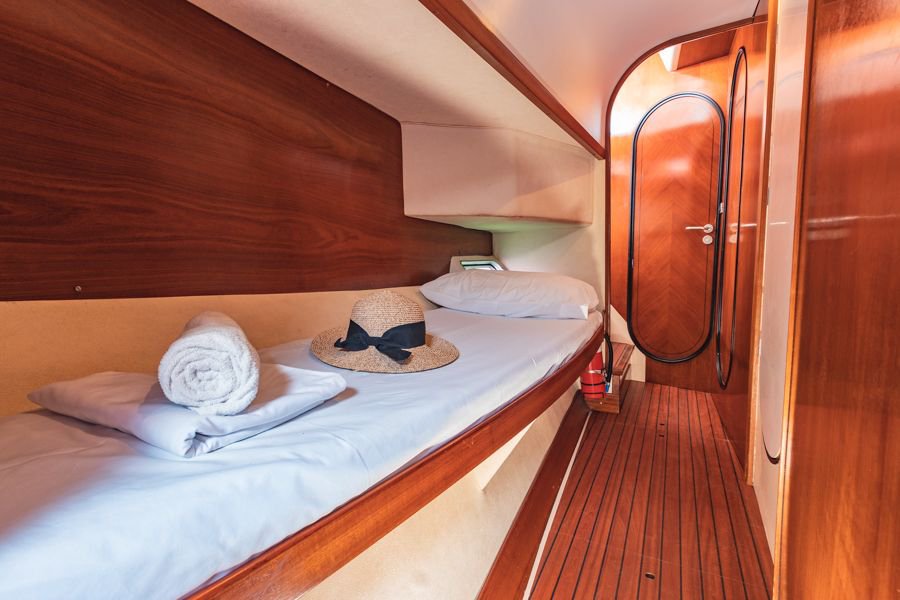 Interior of a sailing yacht cabin featuring a neatly made single bunk bed with a towel and straw hat, polished timber finishes, and a private ensuite door