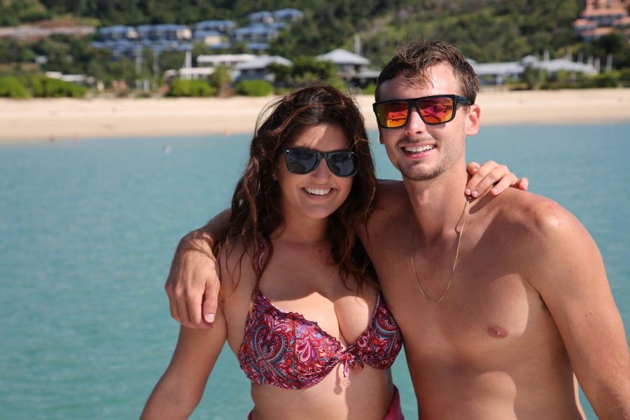 Smiling couple wearing sunglasses standing together by turquoise waters with a sandy beach and hillside in the background in the Whitsundays