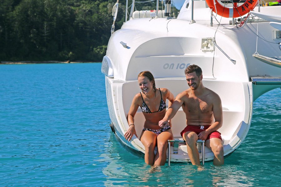 Young couple sitting on the back swim platform of a catamaran with their feet in clear turquoise water in the Whitsundays.