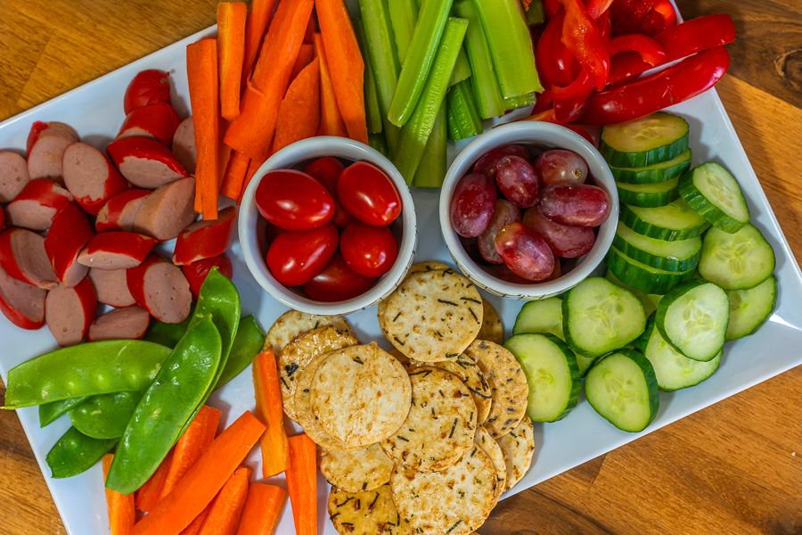 Overhead view of a grazing platter with sliced cucumber, carrot sticks, celery, cherry tomatoes, grapes, crackers and sliced sausage
