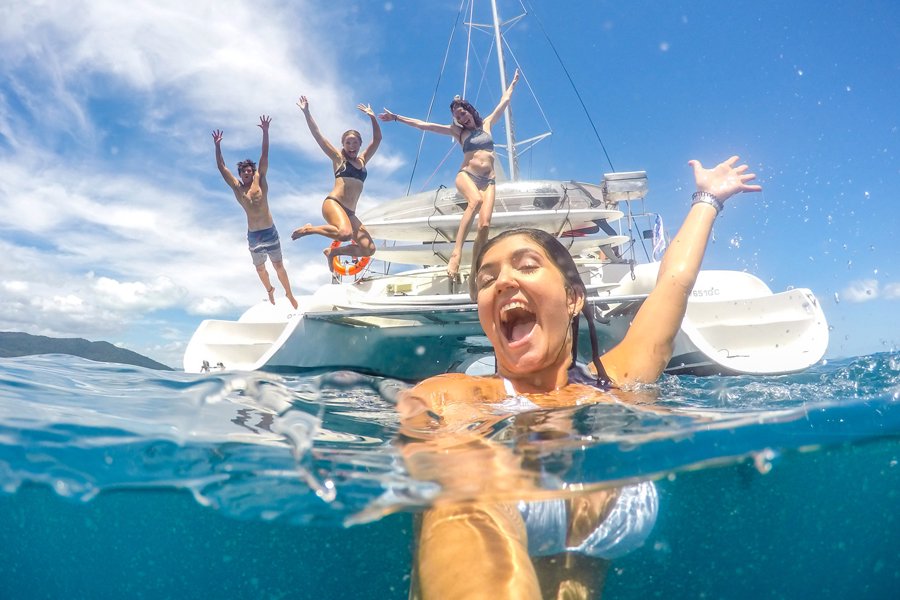 Group of friends jumping off a sailing catamaran into clear turquoise water in the Whitsundays, with one woman smiling in the foreground.