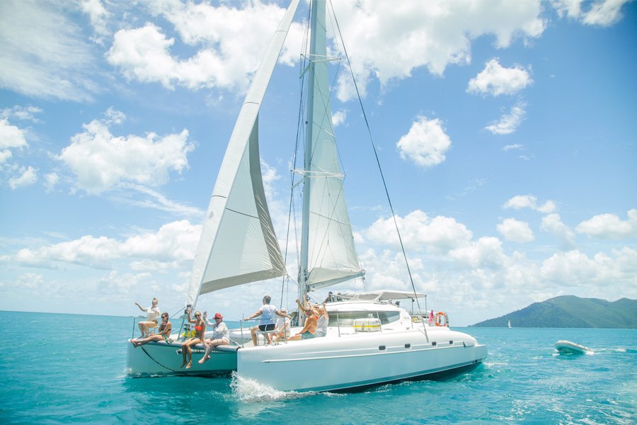 Friends relaxing and enjoying the sunshine on a sailing catamaran cruising through the turquoise waters of the Whitsundays, Queensland.