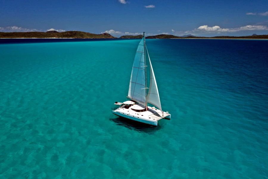 Aerial view of a white sailing catamaran cruising across clear turquoise waters in the Whitsundays, Queensland, Australia.