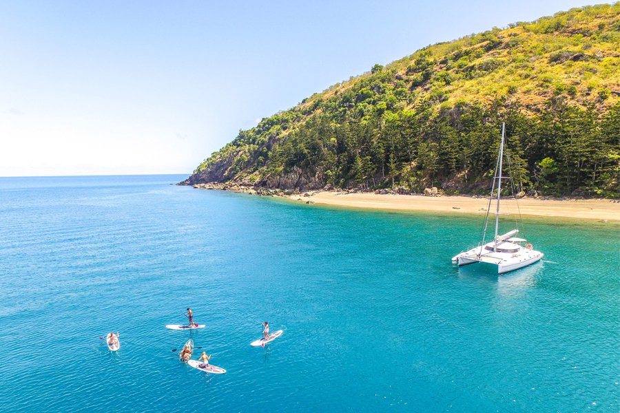 Group of people paddleboarding on turquoise water near a white catamaran anchored off a secluded Whitsundays beach