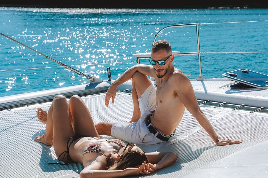 Couple sunbathing on the front deck net of a sailing catamaran in the Whitsundays, surrounded by sparkling turquoise water
