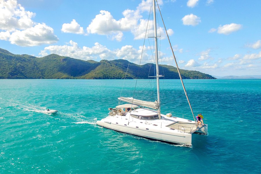 A white sailing catamaran cruising through turquoise waters in the Whitsundays with lush green islands in the background under a blue sky