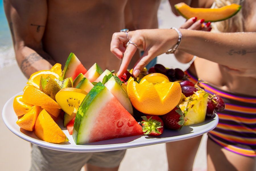 Close-up of a colourful tropical fruit platter with watermelon, orange slices, kiwi, grapes, strawberries and pineapple being shared by guests on a sunny beach