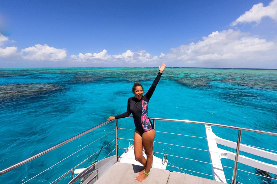 Woman in a long-sleeve swimsuit standing on the deck of a boat with her arm raised, overlooking the bright turquoise waters of the Great Barrier Reef in the Whitsundays
