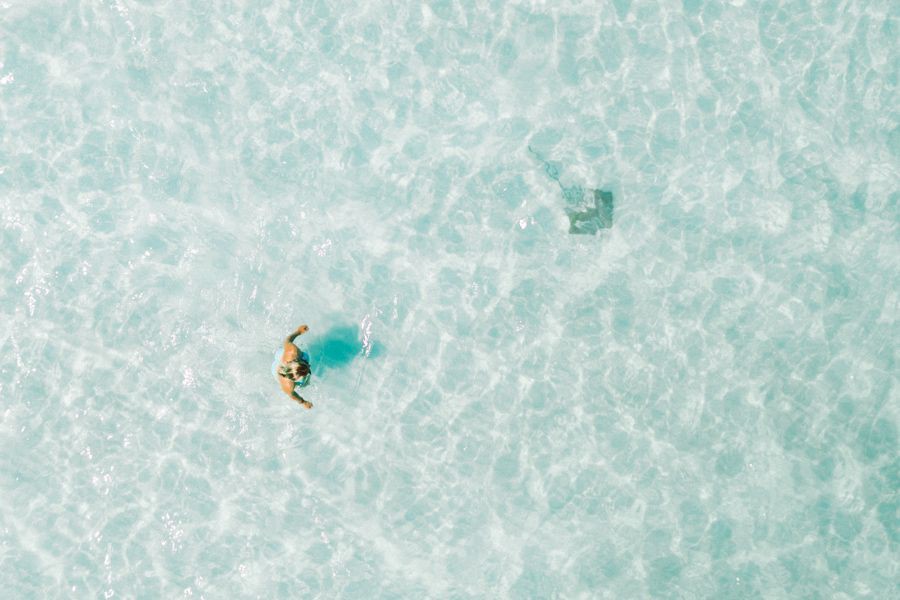 Top-down aerial view of a person wading through shallow, crystal-clear turquoise water in the Whitsundays with rippled white sand visible beneath the surface
