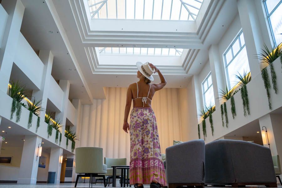 Woman in a sunhat and floral maxi skirt standing in a bright, modern resort lobby with high ceilings, skylights, and indoor greenery on Hamilton Island