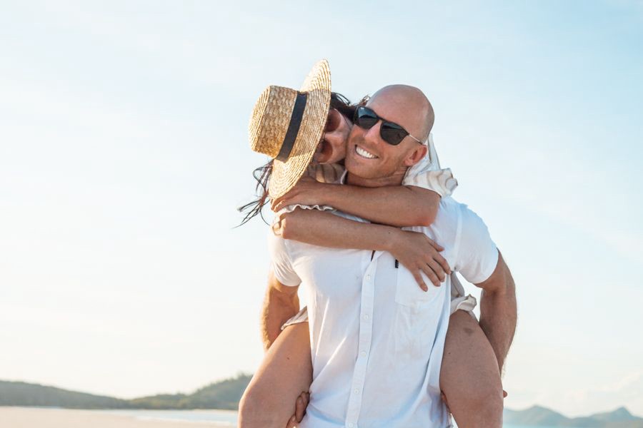 Smiling couple enjoying a playful piggyback moment on a sunny beach in the Whitsundays, with clear skies and ocean in the background