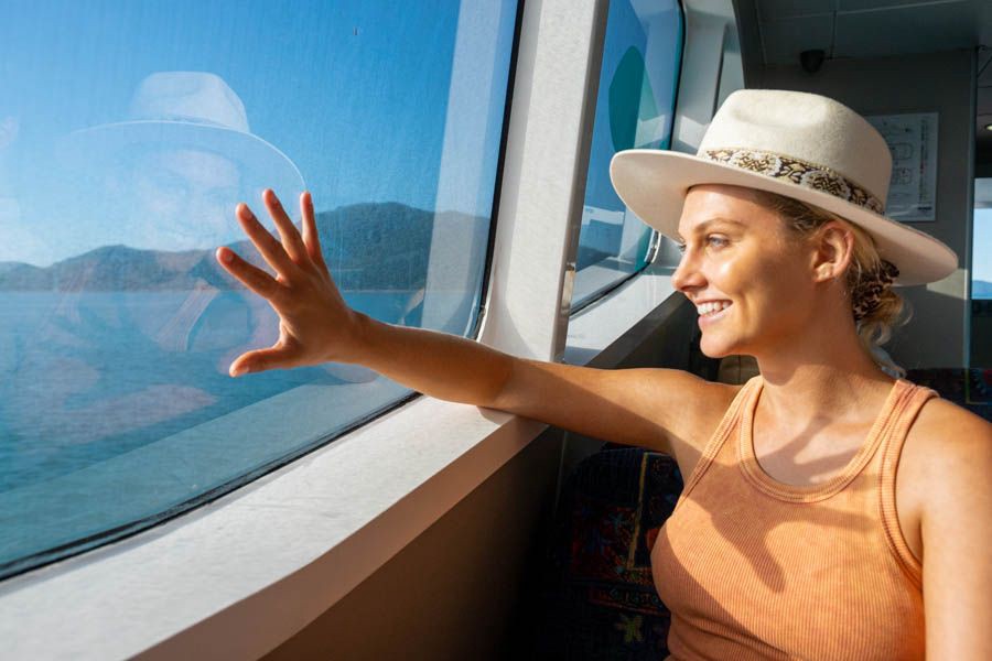 Woman wearing a sun hat smiling and looking out the window of a ferry at the turquoise waters and island scenery in the Whitsundays