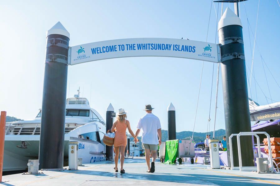 Couple holding hands walking along a marina jetty beneath a “Welcome to the Whitsunday Islands” sign, with boats and yachts docked on either side