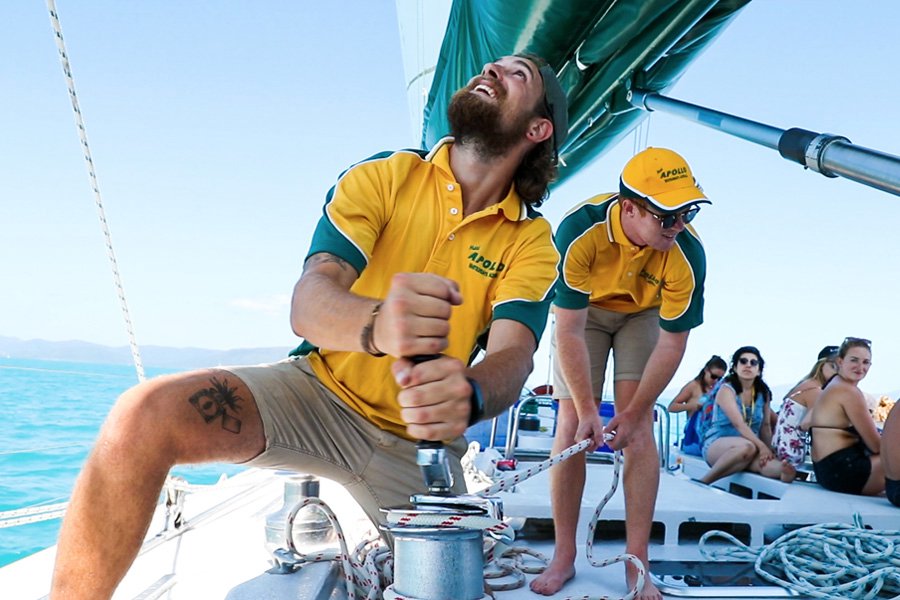 Two Apollo crew members adjusting sails on a catamaran while guests relax on deck in the Whitsundays