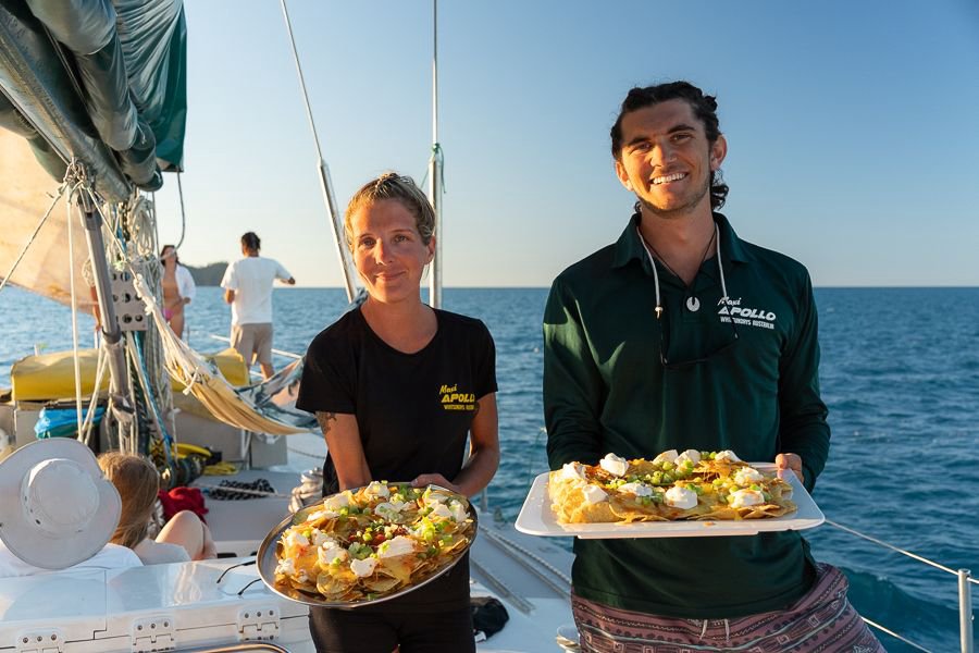 Maxi Apollo crew members serving platters of nachos on deck during a sunset sail in the Whitsundays