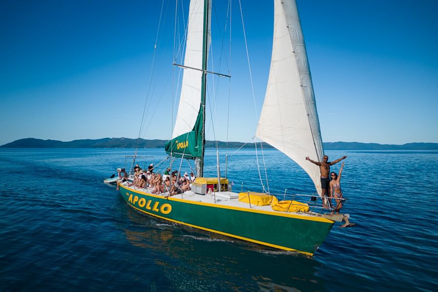 Guests relaxing and posing on the deck of the green and yellow Apollo sailing yacht cruising through calm blue waters in the Whitsundays.
