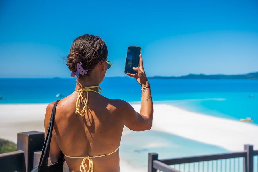 Woman in a yellow bikini top taking a photo with her phone at Hill Inlet Lookout, overlooking the swirling white sands and turquoise waters of Whitehaven Beach in the Whitsundays