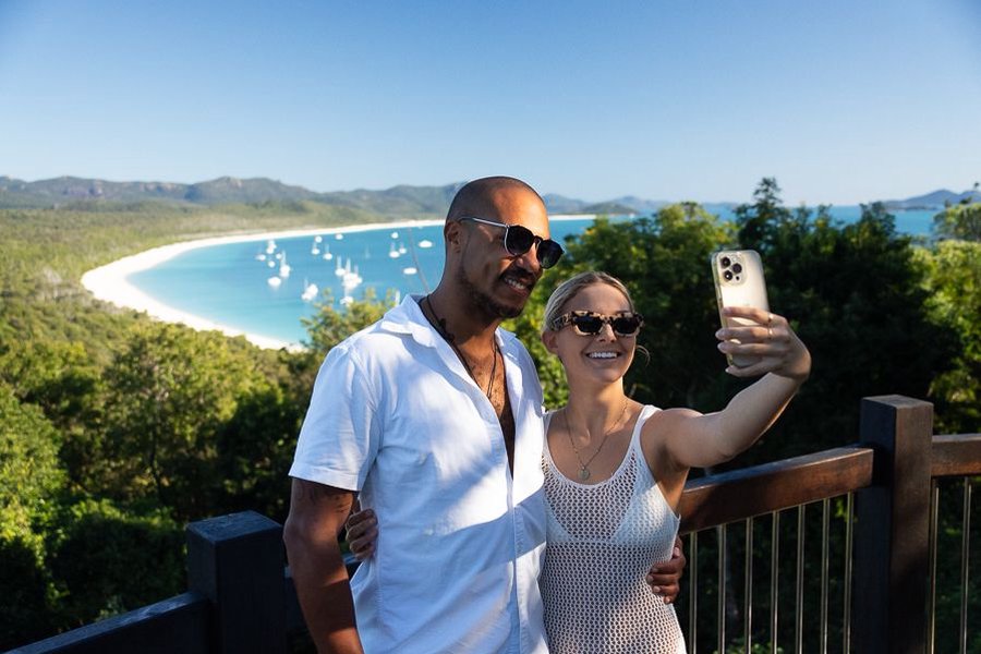 Smiling couple taking a selfie at Hill Inlet lookout with Whitehaven Beach and anchored boats in the background, Whitsundays