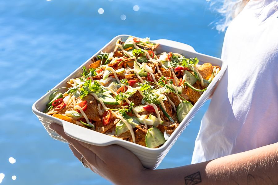 Tray of freshly prepared nachos topped with vegetables and sauce held on a boat with sparkling ocean in the background