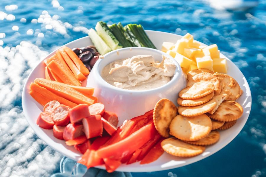 Close-up of a grazing platter with hummus, crackers, cheese cubes, sliced vegetables and olives held above sparkling turquoise ocean water during a Whitsundays sailing trip