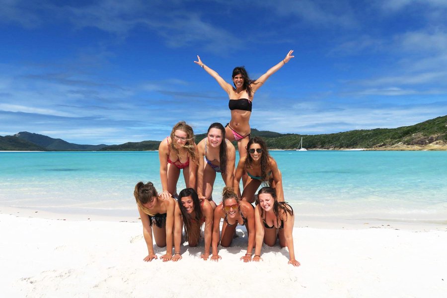 Group of friends in bikinis forming a human pyramid on the white sands of Whitehaven Beach with turquoise Whitsundays waters behind them