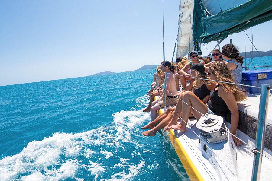 Group of young travellers sitting along the side of a sailing yacht with their feet over the edge, cruising through bright turquoise waters in the Whitsundays on a sunny day
