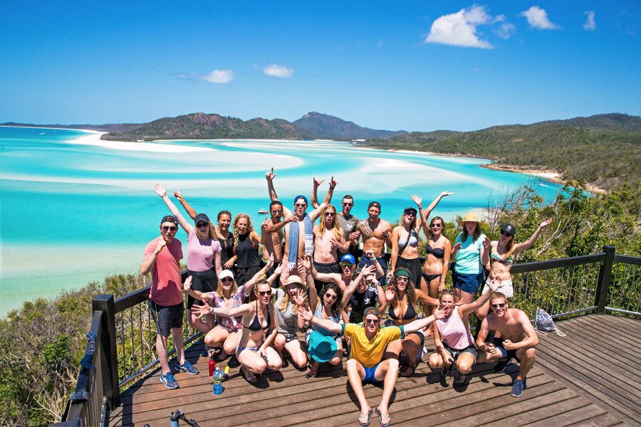 Large group of friends posing with arms raised at Hill Inlet Lookout overlooking the turquoise waters and white sand swirls of Whitehaven Beach