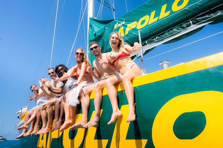 Group of smiling young adults sitting along the side rail of the green and yellow Maxi Apollo sailboat with legs over the edge, enjoying a sunny day sailing in the Whitsundays