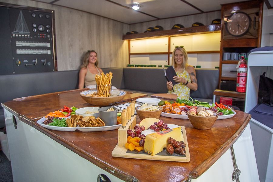 Guests seated in a yacht saloon with a large wooden table set with cheese boards, crackers, fresh fruit and antipasto platters