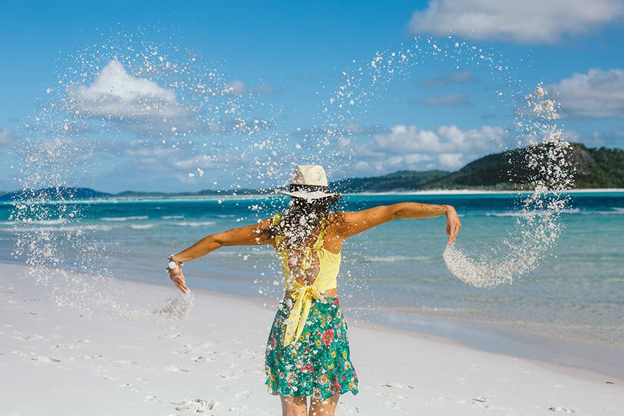 Woman in a yellow top and floral skirt tossing fine white sand into the air on Whitehaven Beach, with turquoise water and island hills in the background