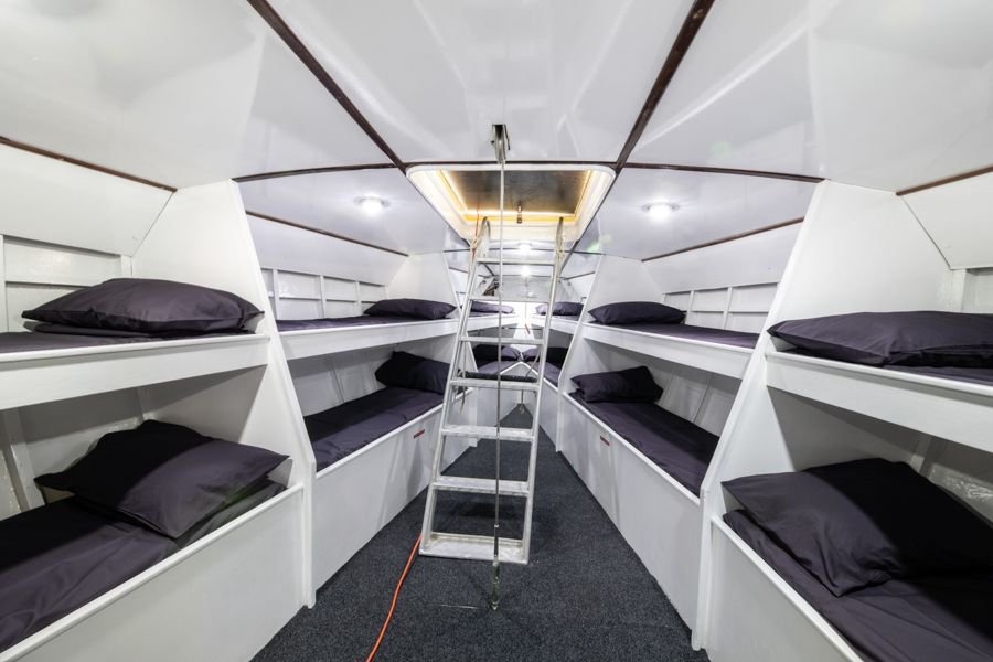 Interior of the Maxi Apollo sailboat featuring shared bunk-style cabin accommodation with multiple single berths, dark bedding, and a central ladder leading to the deck above
