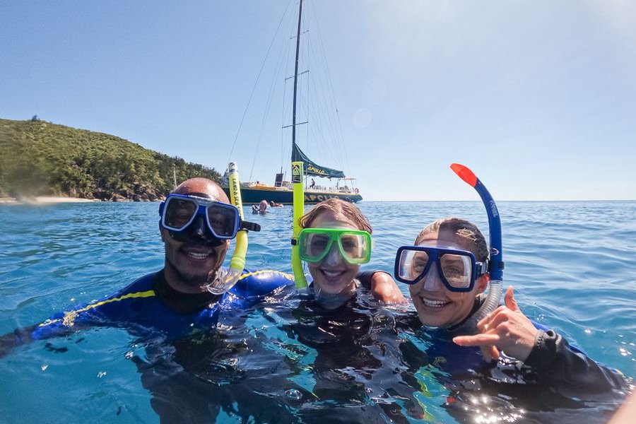Three friends smiling in the ocean wearing snorkel masks and snorkels with a sailing boat anchored behind them in the Whitsundays