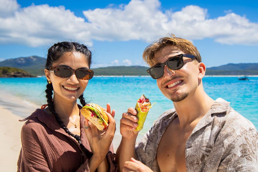 Smiling couple holding fresh wraps while standing on a white sandy beach with turquoise water in the Whitsundays, Queensland.