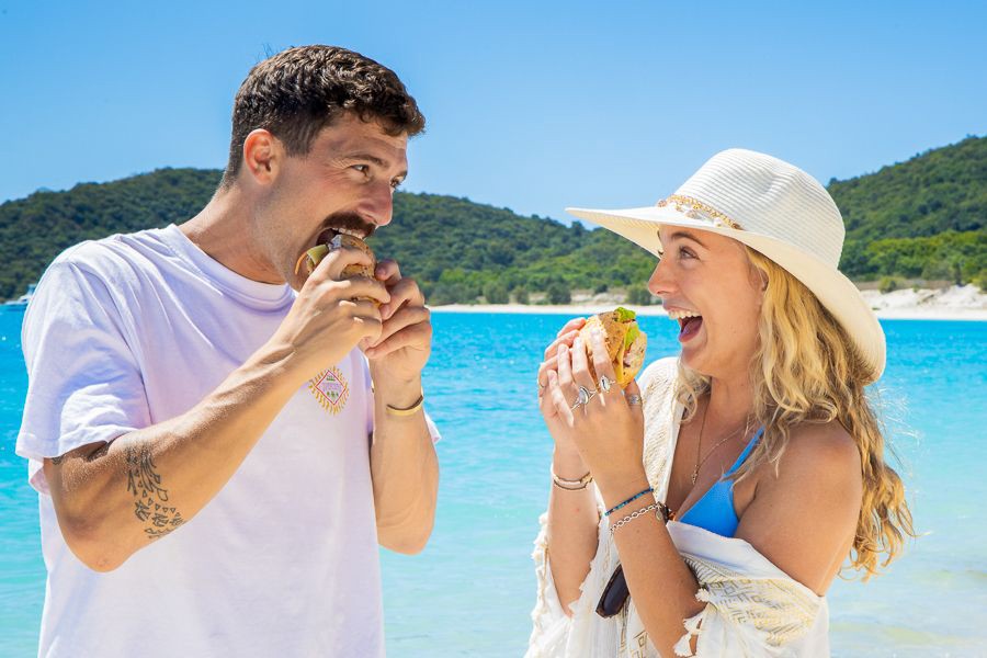 Couple laughing and eating burgers on Whitehaven Beach with turquoise water and tropical island backdrop in the Whitsundays, Queensland.