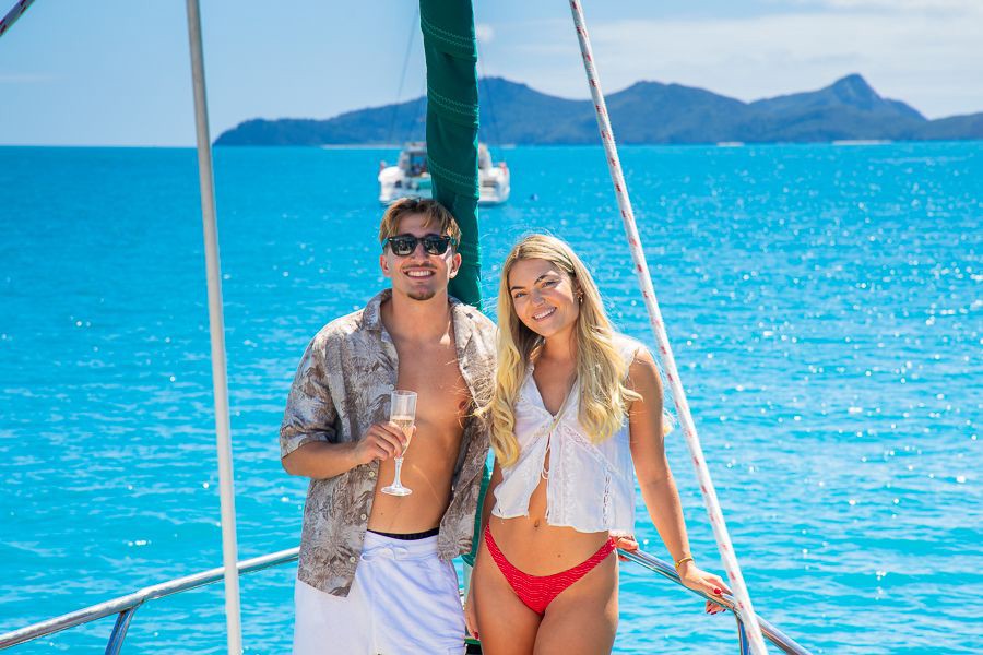 Smiling couple standing on the deck of a sailboat with turquoise Whitsundays water and island mountains in the background, Queensland, Australia.