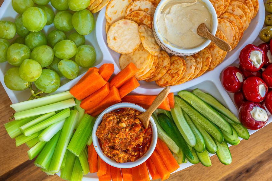 Colourful grazing platter featuring crackers, hummus, sundried tomato dip, fresh grapes, carrot and celery sticks, cucumber slices, and stuffed peppers.