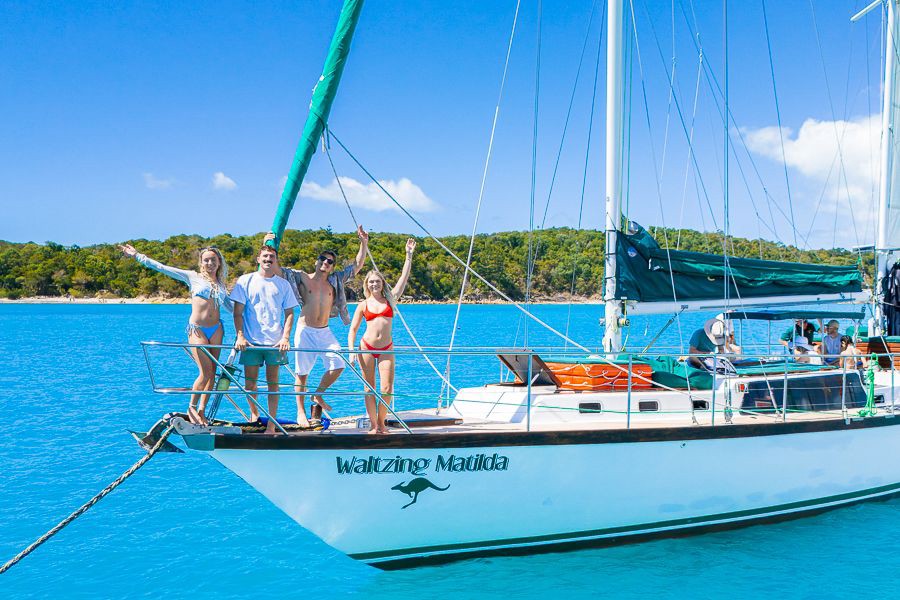 Group of friends standing at the bow of the sailing yacht Waltzing Matilda, smiling and waving while anchored in bright turquoise waters near a lush island in the Whitsundays