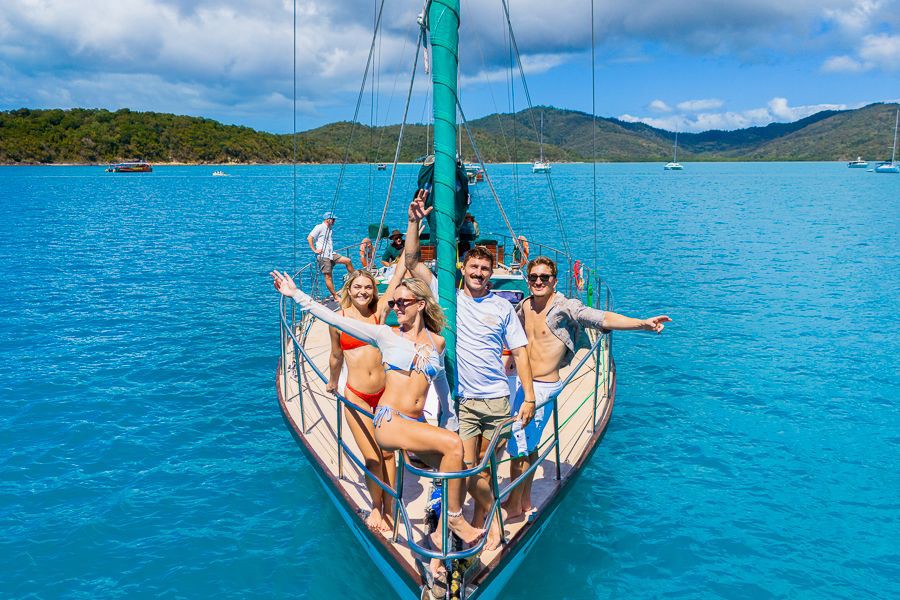 Group of friends posing at the bow of a sailboat surrounded by turquoise water and island scenery in the Whitsundays, Australia.