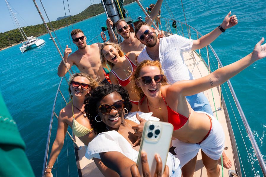 Group of friends in swimwear smiling and taking a selfie on the deck of a sailing yacht surrounded by turquoise waters and island scenery in the Whitsundays