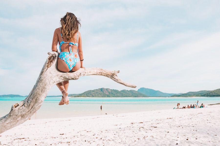 Woman sitting on a driftwood tree overlooking the white silica sands and turquoise waters of Whitehaven Beach in the Whitsundays, Queensland.