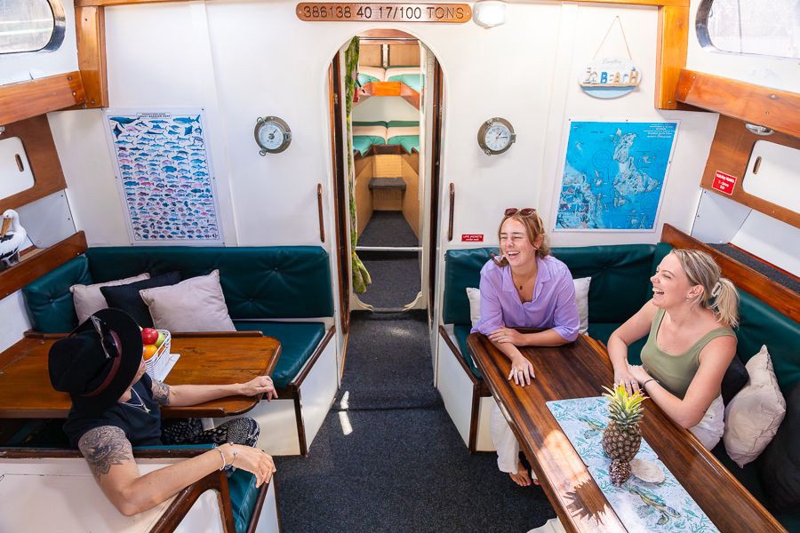 Guests relaxing inside the Waltzing Matilda yacht cabin with cushioned seating and wooden tables during a Whitsundays sailing tour