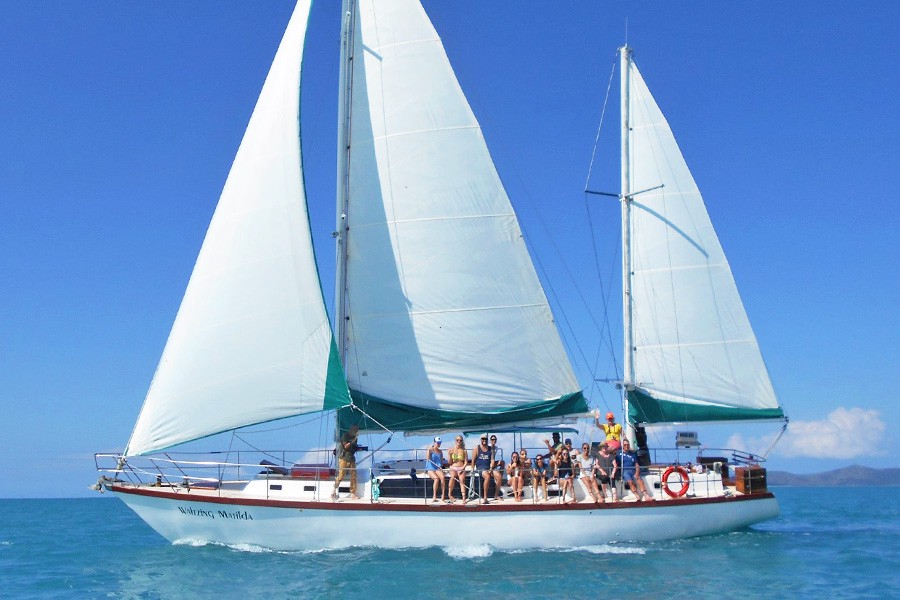 Group of guests relaxing on the deck of a large white sailing yacht with full sails raised, cruising through turquoise waters in the Whitsundays