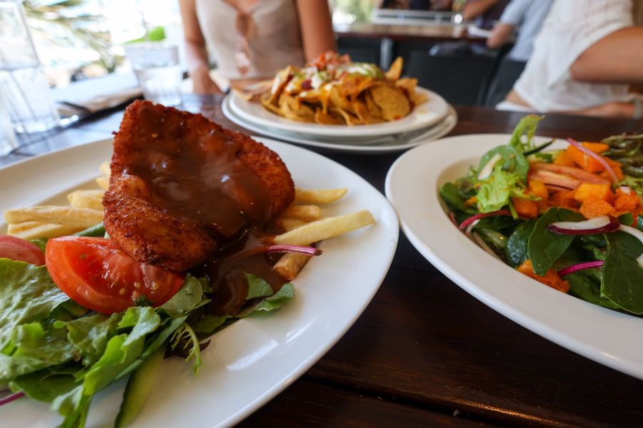 Plates of schnitzel with chips and salad, fresh garden salad, and loaded nachos served at a marina-side restaurant