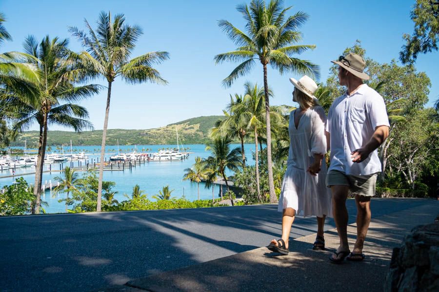 Couple walking hand in hand along a palm-lined road overlooking Hamilton Island Marina in the Whitsundays