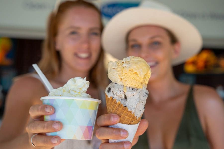 Two women holding ice cream cone and cup with whipped cream, smiling at a beachside shop