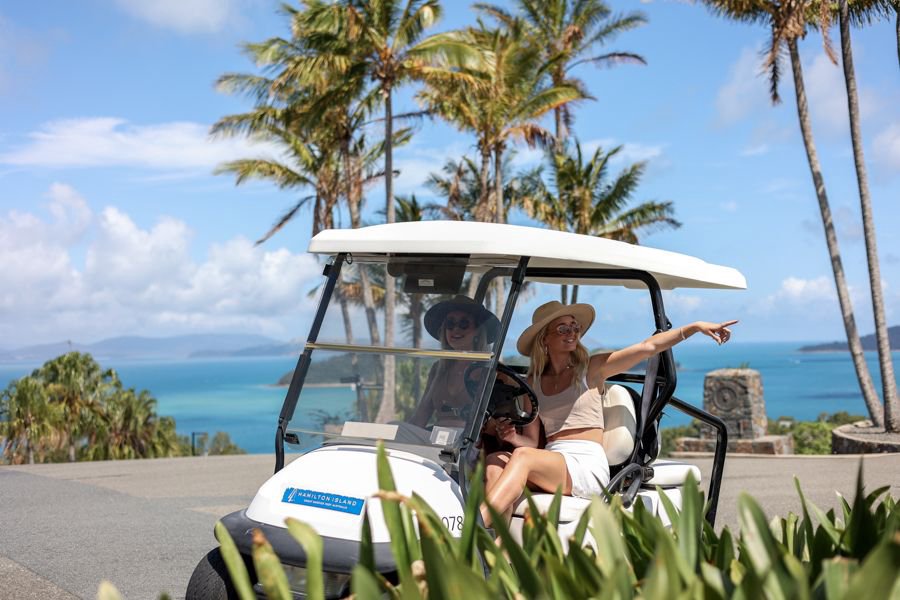 Two women riding in a golf buggy on Hamilton Island with palm trees and turquoise ocean in the background