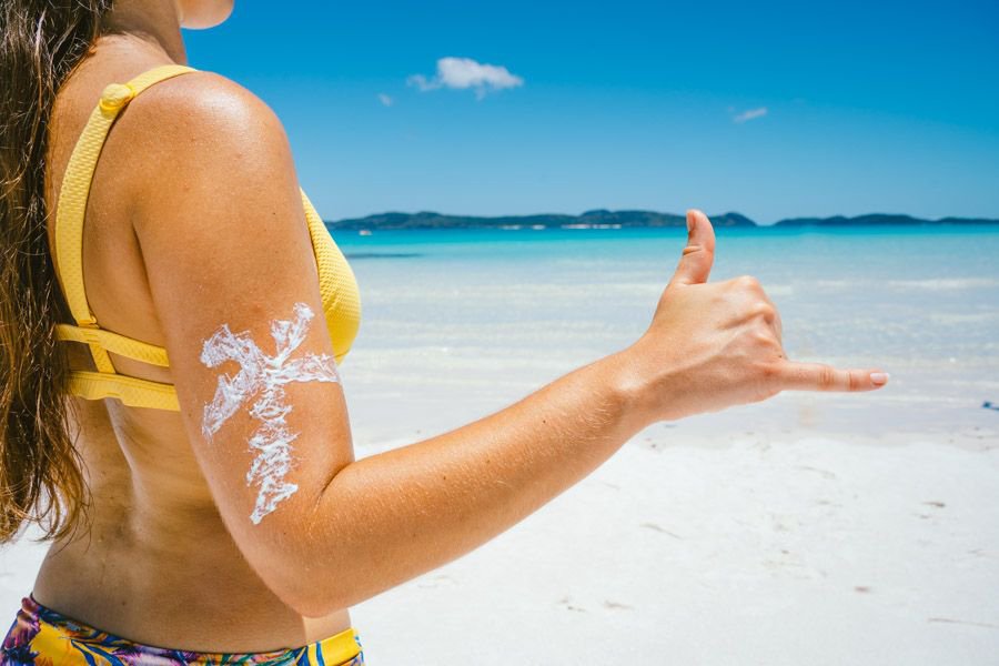 Close-up of a woman in a yellow bikini at Whitehaven Beach applying sunscreen on her arm while making a shaka hand gesture, with turquoise ocean and islands in the background
