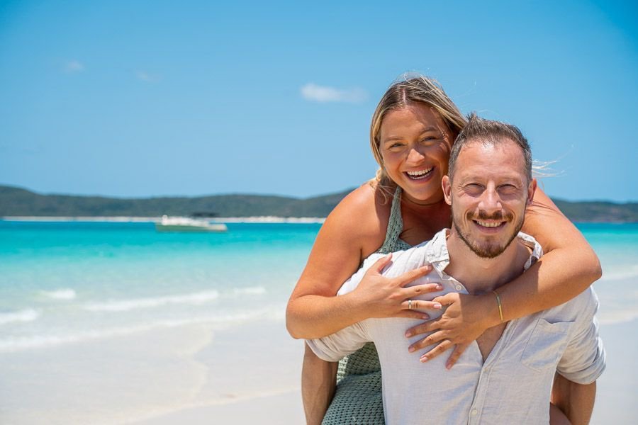 Smiling couple at Whitehaven Beach with the woman giving the man a piggyback, turquoise ocean and island hills in the background