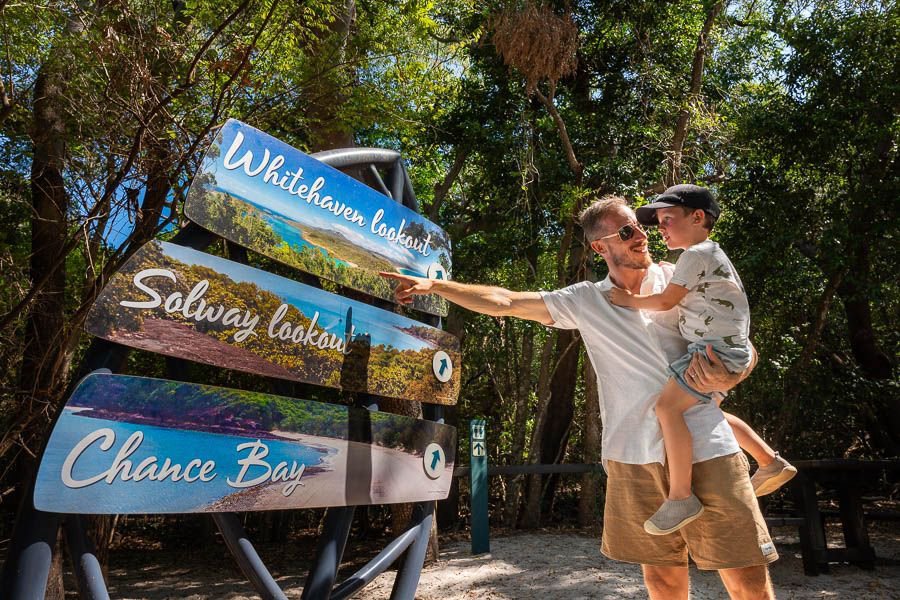 Father holding his young son while pointing at a signpost for Whitehaven Lookout, Solway Lookout and Chance Bay in a tropical forest setting