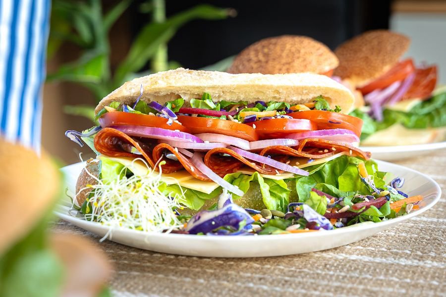 A freshly made gourmet sandwich filled with salami, cheese, tomato, red onion, and leafy greens, served on a plate with a side salad